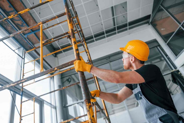 The man was dressed in grey work attire, black T-shirt and orange helmet and work gloves. He is holding an orange scaffolding by its base, which is partially stripped of paint.