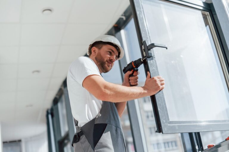 A man in grey construction garb, installs double-glazed windows. The windows have white frames and the weather outside the window is sunny.