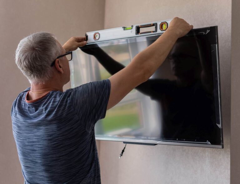 A man mounts a television on a wall. To keep it straight, he uses a spirit level, which is placed on top of the TV frame.