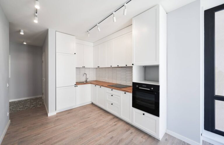 Modern white kitchen in the form of a kitchenette. White cabinets and appliances are visible. There is spot lighting on the ceiling. This kitchen design looks costly.